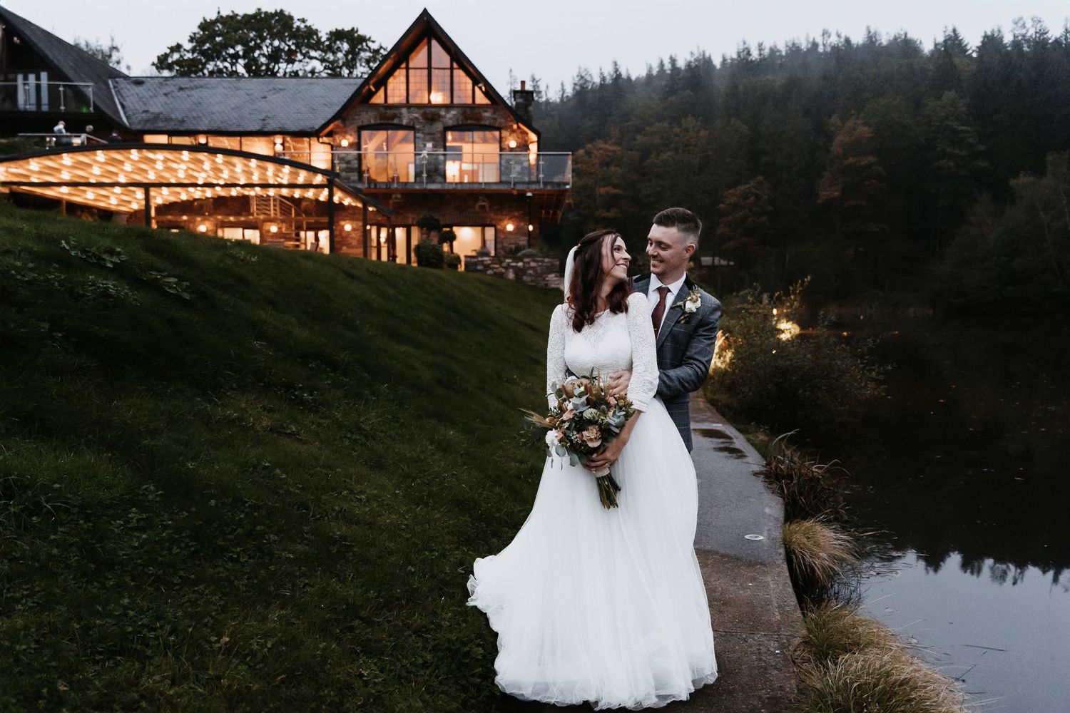 Wedding couple by illuminated rustic lodge overlooking misty pond at dusk.