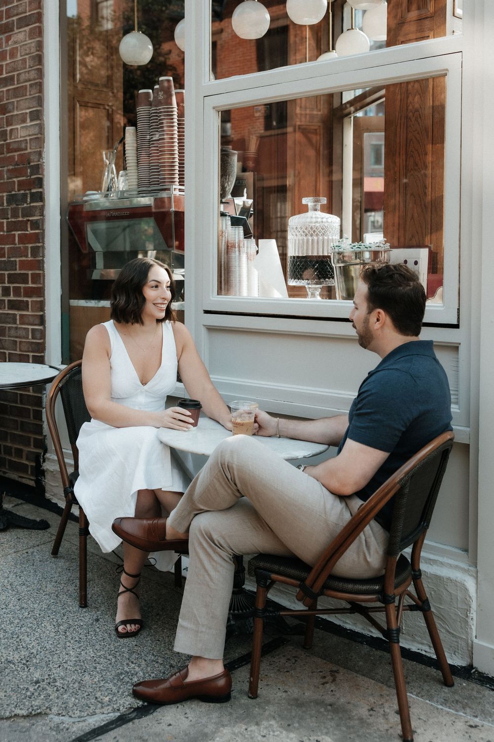 A couple sits together on a wooden bench outside a brick building decorated with white paper lanterns.