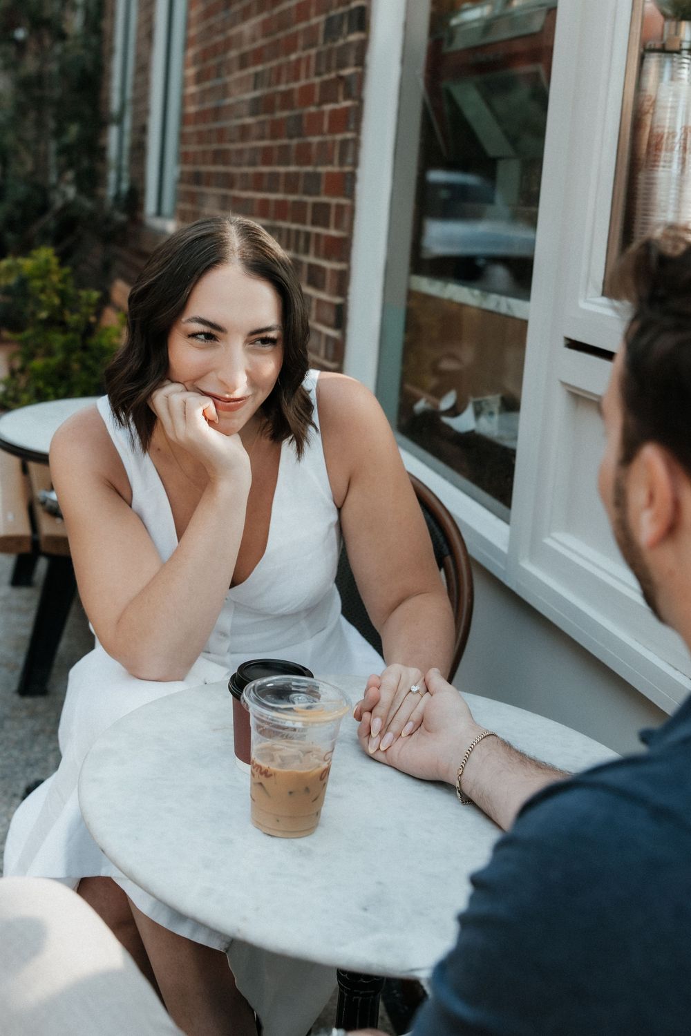 Two people sitting at an outdoor cafe table with iced coffee drinks on a sunny day near a brick building.