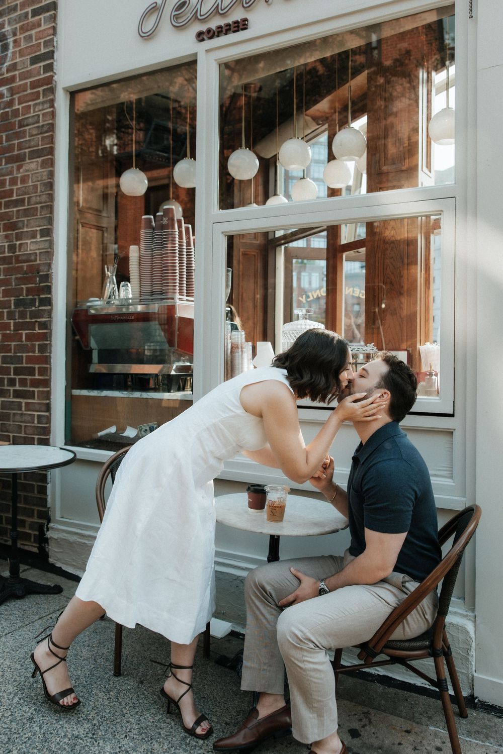 A romantic photo series showing a couple sharing a playful kiss while seated outside a cafe with brick walls.