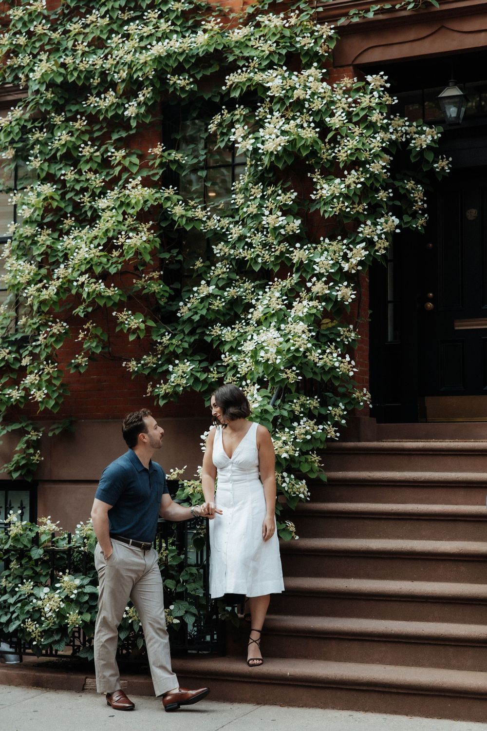 Two people sharing a tender moment on brick steps framed by lush greenery and white flowers.