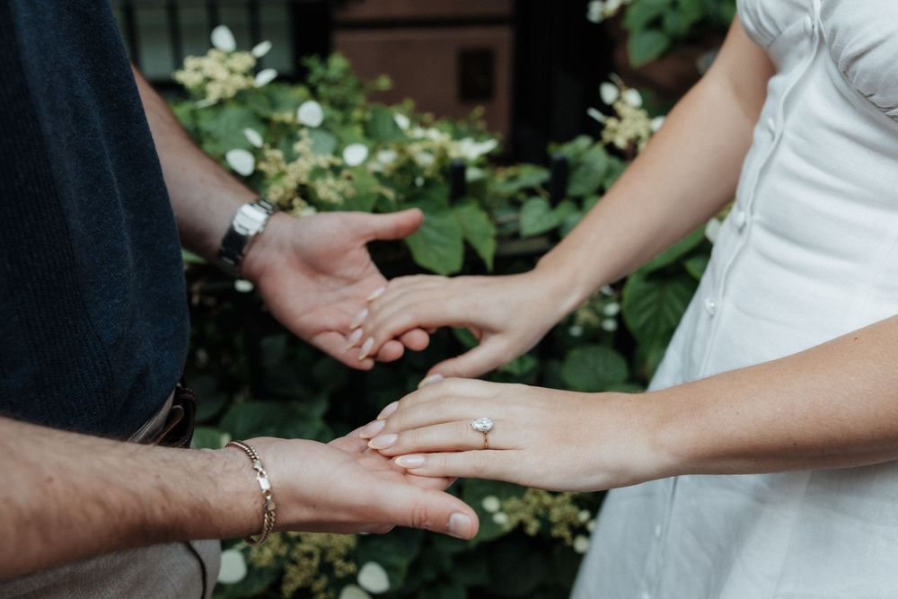 Close up view of hands exchanging wedding rings during an outdoor wedding ceremony with greenery in background.