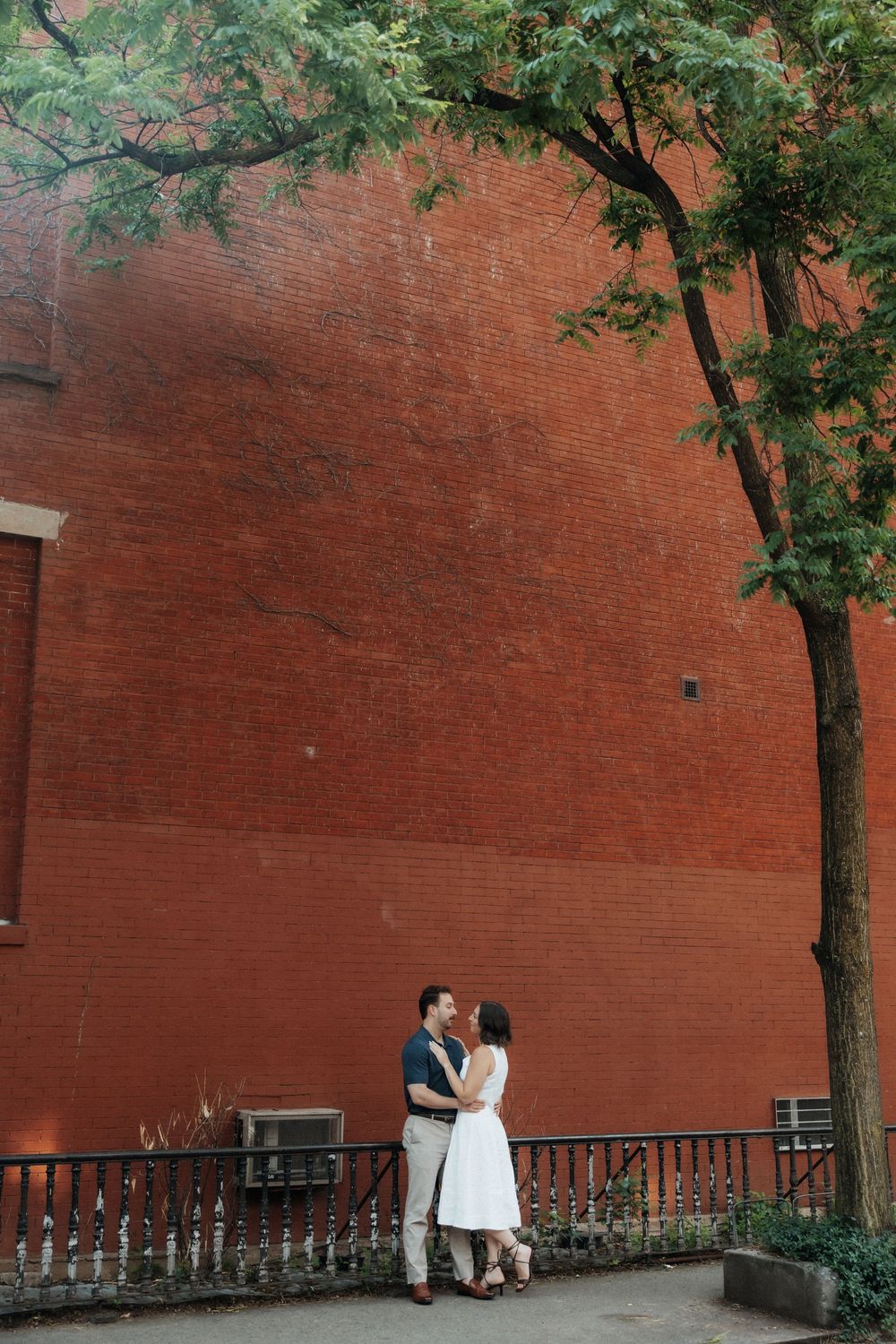 A couple stands together against a rustic red brick wall with leafy trees providing natural shade.