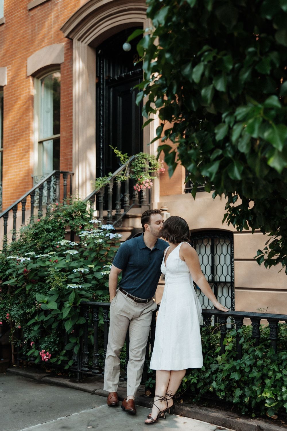 Four photos show a couple walking and peeking through a window of a historic brownstone building on a leafy street.