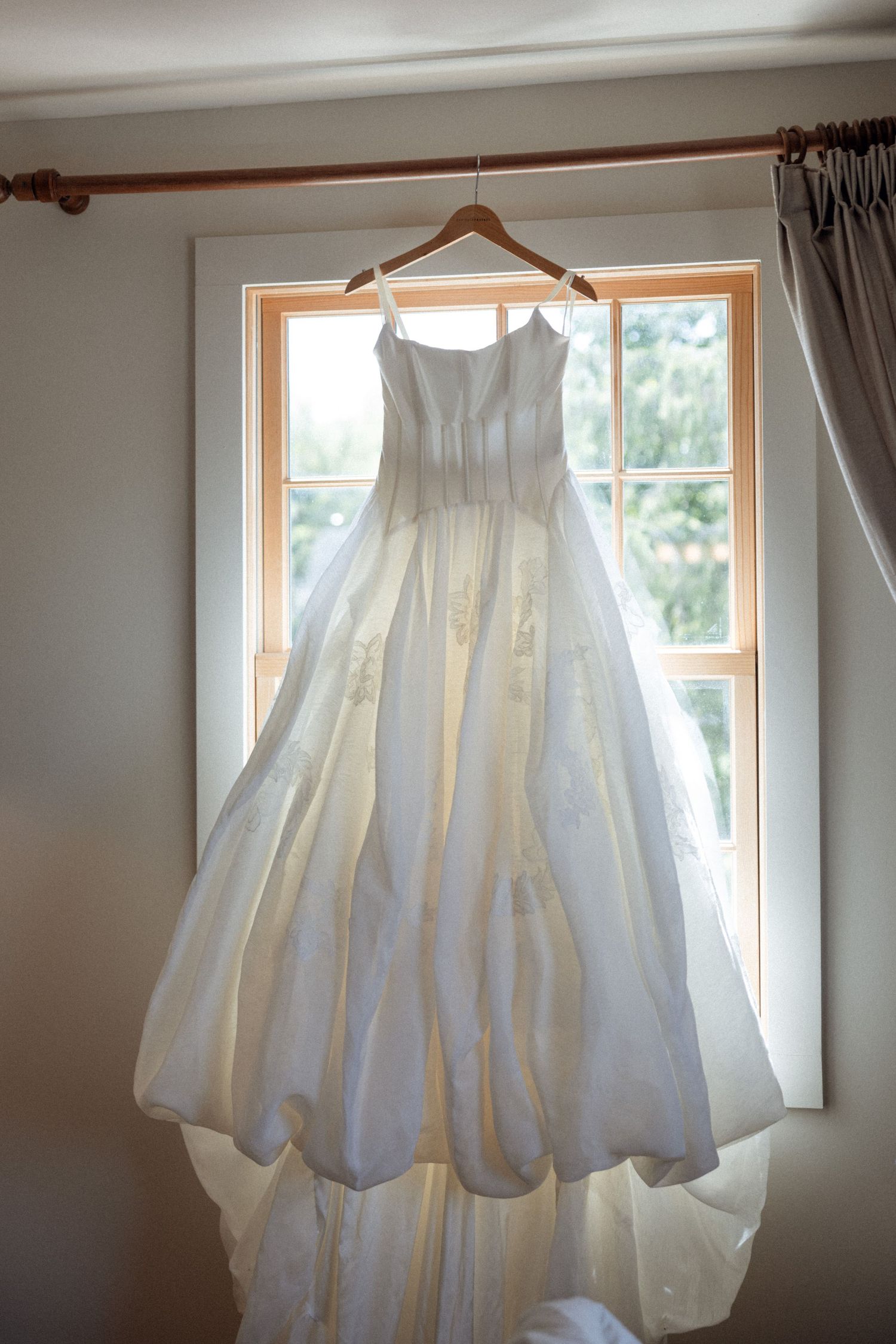 A white wedding dress hangs in front of windows, backlit by natural daylight.