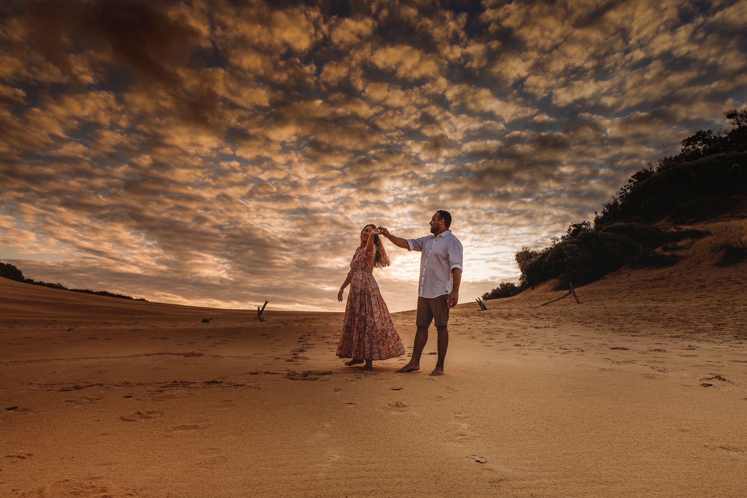 A couple walks together on a sandy beach under dramatic orange clouds at sunset.