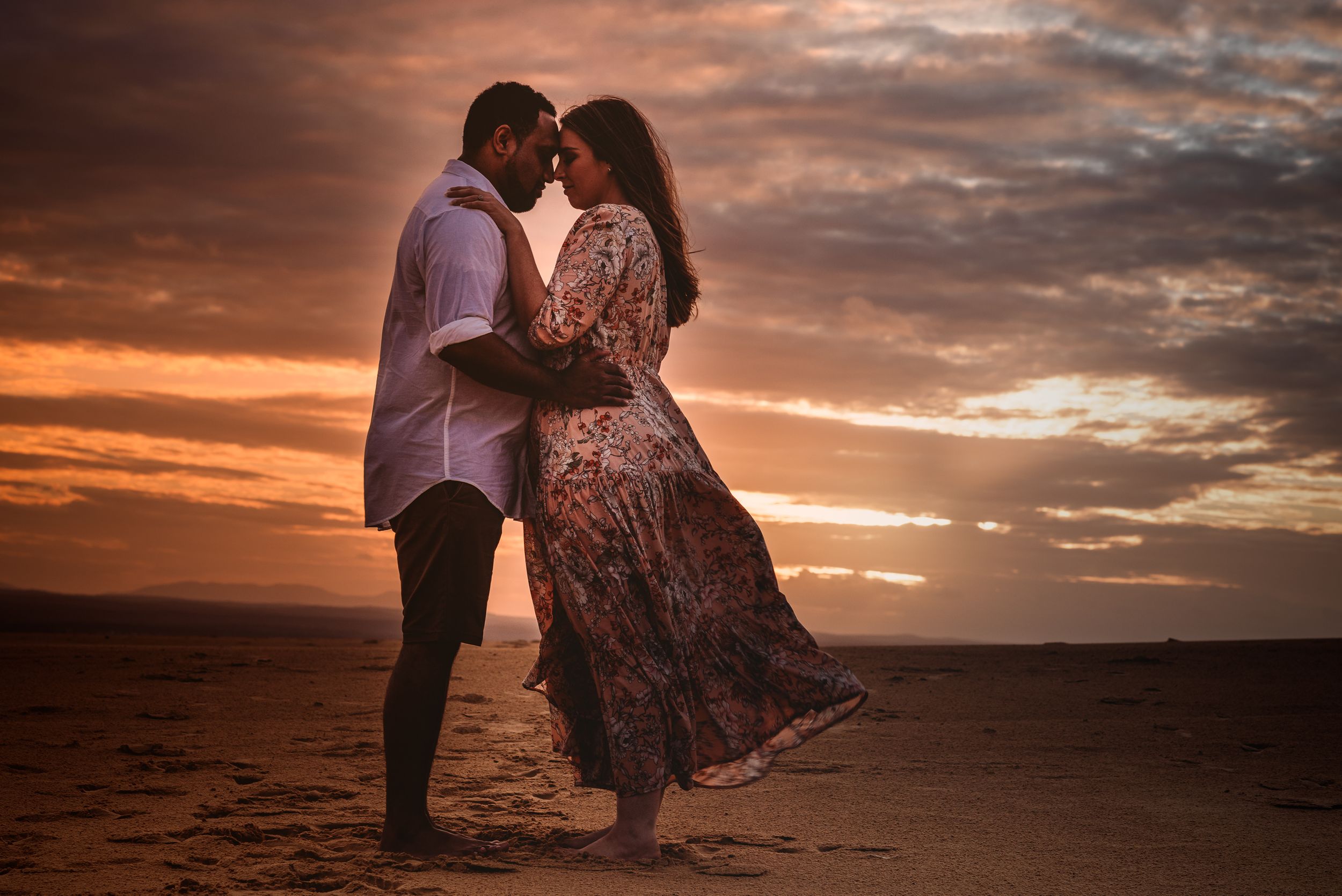 A romantic couple embraces on a beach during a dramatic sunset with orange cloudy skies.
