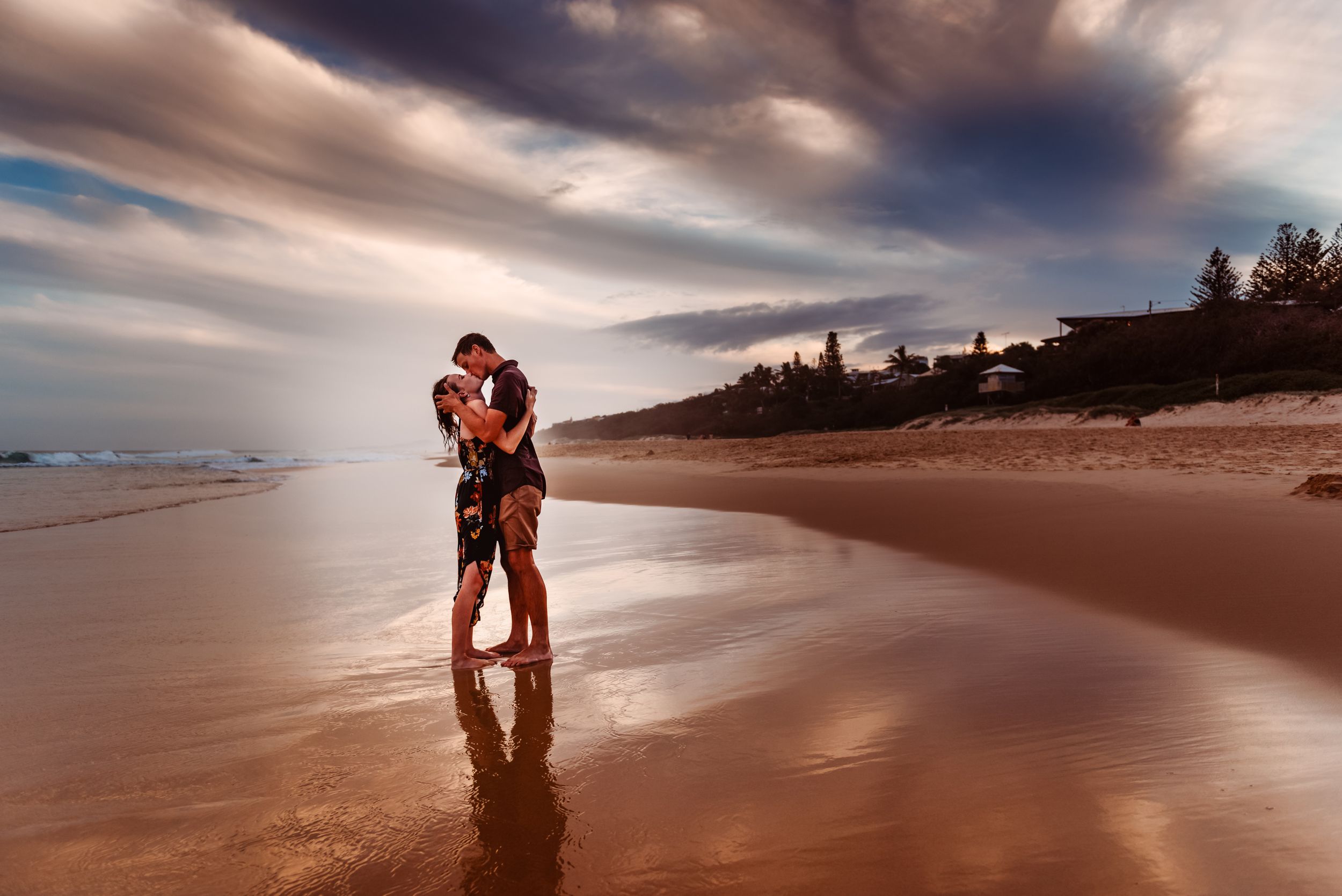 A silhouetted couple sharing an intimate moment on a beach at sunset with dramatic orange clouds.