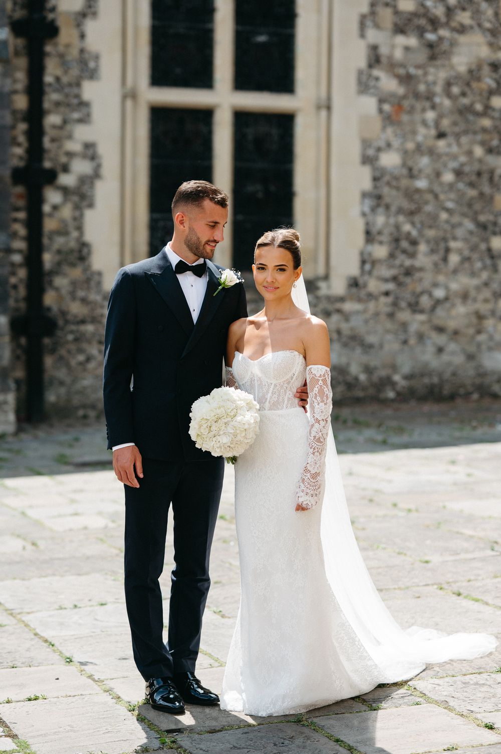 Elegant wedding couple poses for photos against stone building backdrop on sunny day.