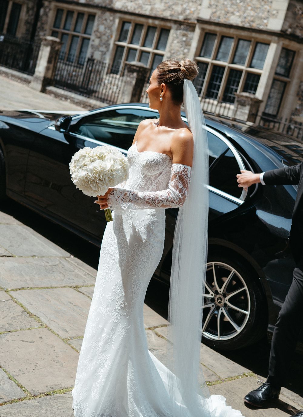 Off-shoulder lace wedding dress with flowing train photographed outside historic stone building.