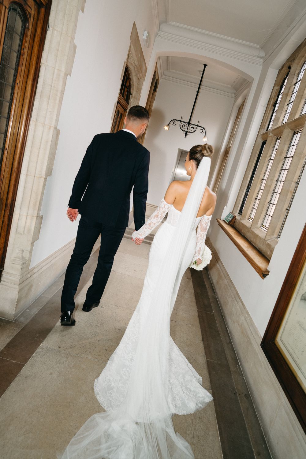 Bride and groom walking down a white corridor with arched windows creating a dramatic wedding photo sequence.