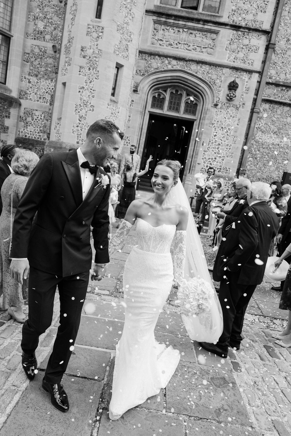A black and white wedding scene with confetti flying as guests celebrate outside a stone building.
