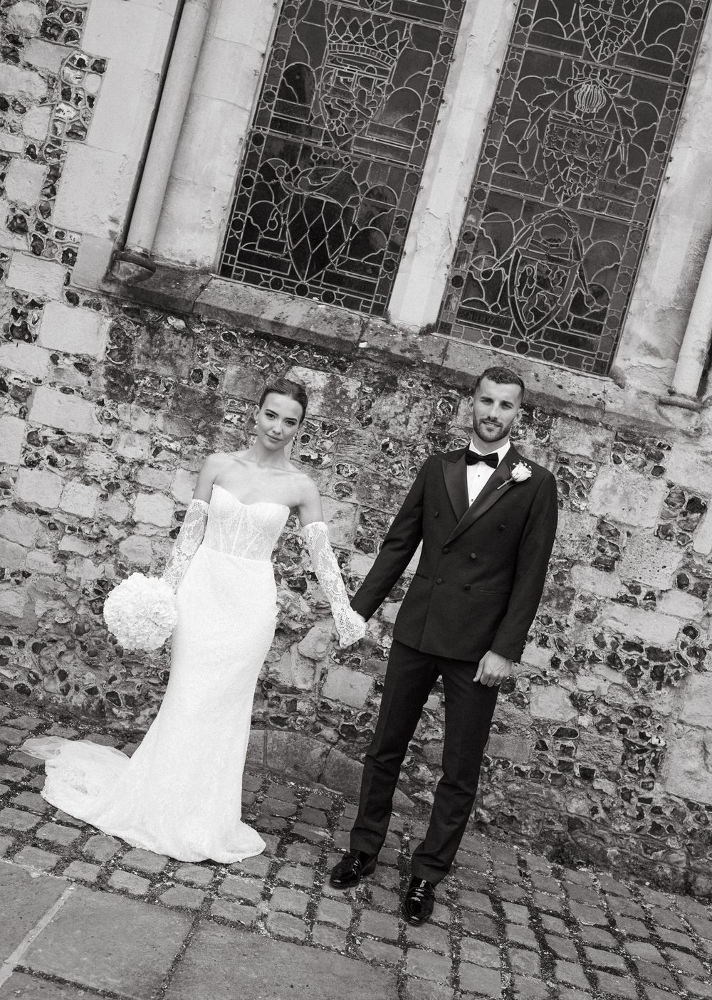 Black and white wedding photo of newlyweds standing together against an ornate stone church wall with stained glass windows.