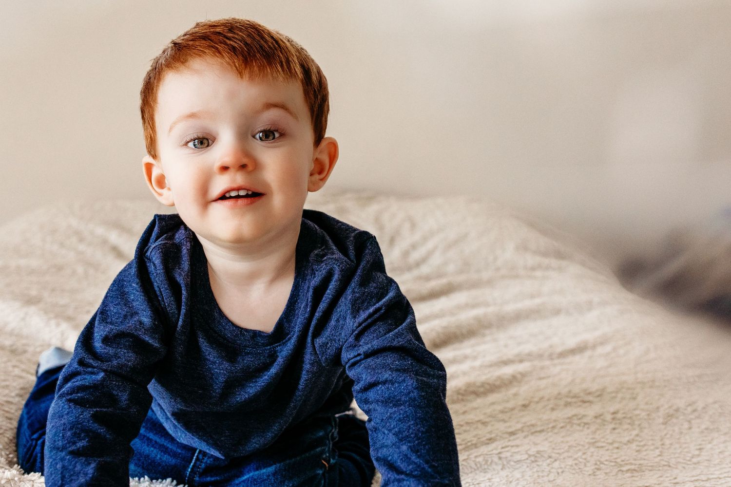 Young toddler in navy sweater sitting on white bed textures while looking off to side with bright expression.