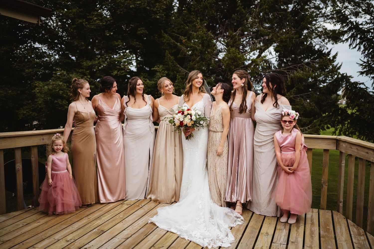 Bridesmaids in pink and nude dresses stand together on wooden deck during an outdoor wedding ceremony.