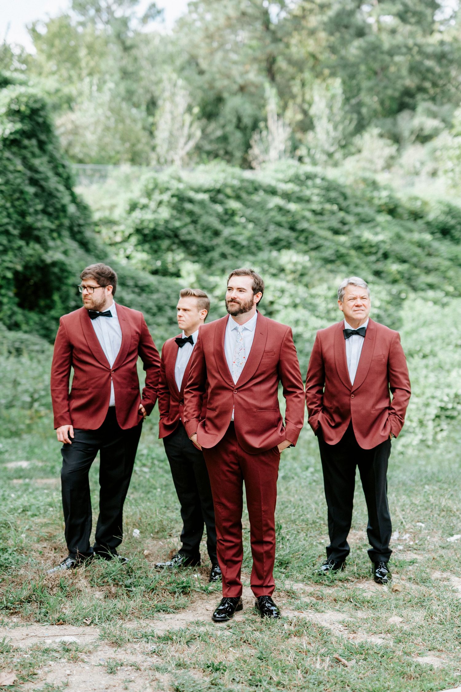 Wedding party in burgundy suits posing outdoors in a coordinated line-up against green foliage.