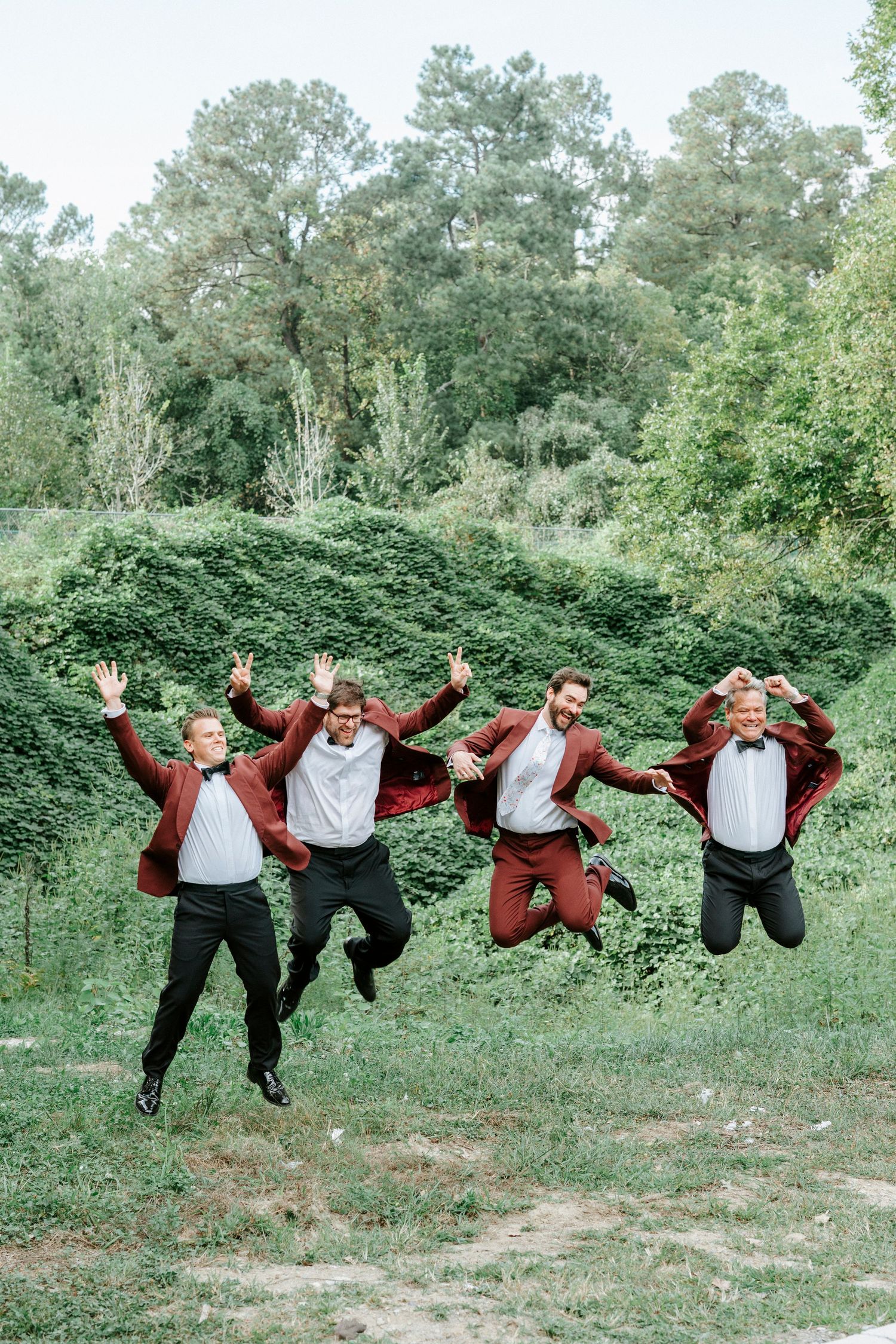 Groomsmen in burgundy suits leaping joyfully in unison outside on a grassy area.
