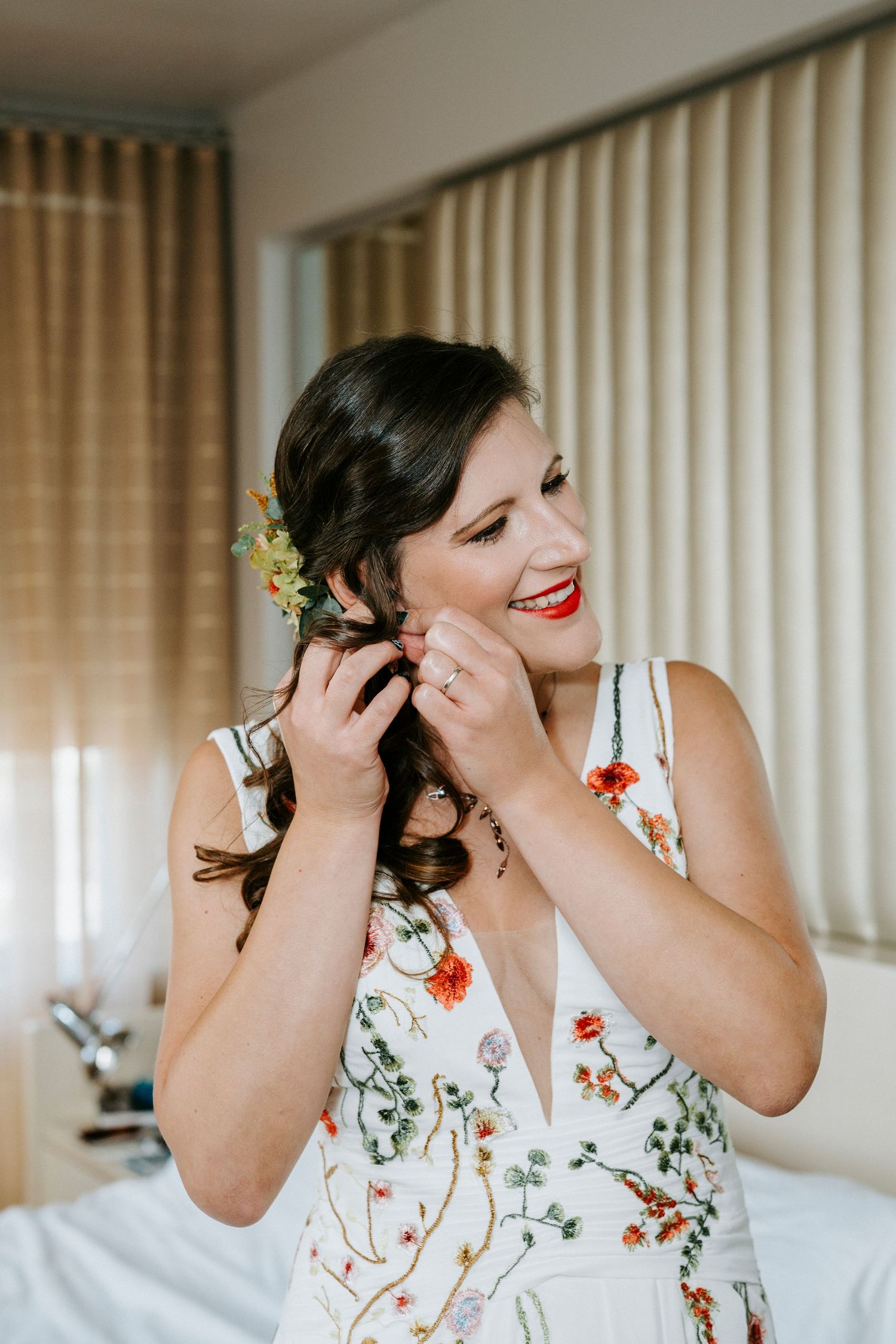 A person in a floral white dress adjusts an earring while smiling and getting ready for a special occasion.