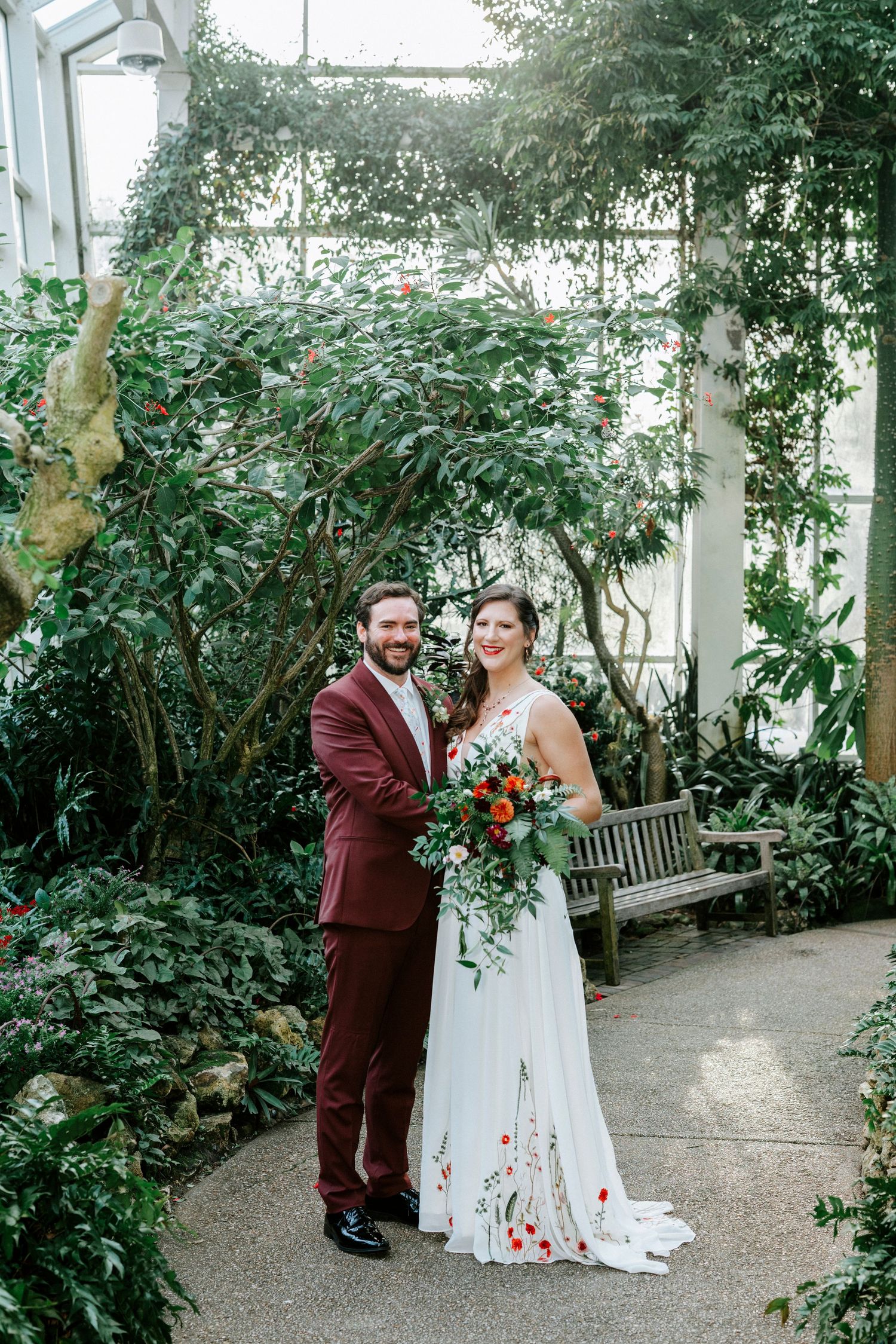 Wedding couple poses in a lush indoor greenhouse conservatory with white and burgundy floral decor.