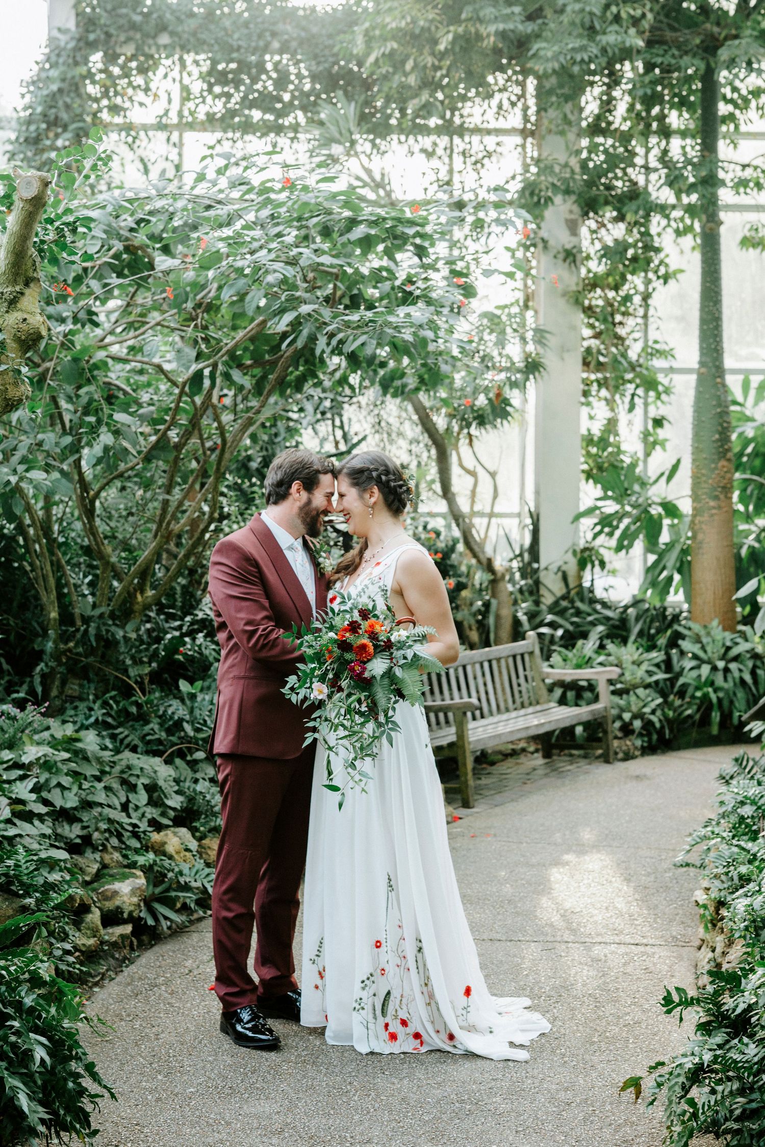 A tender wedding moment between newlyweds on a garden path surrounded by lush greenery and a wooden bench.
