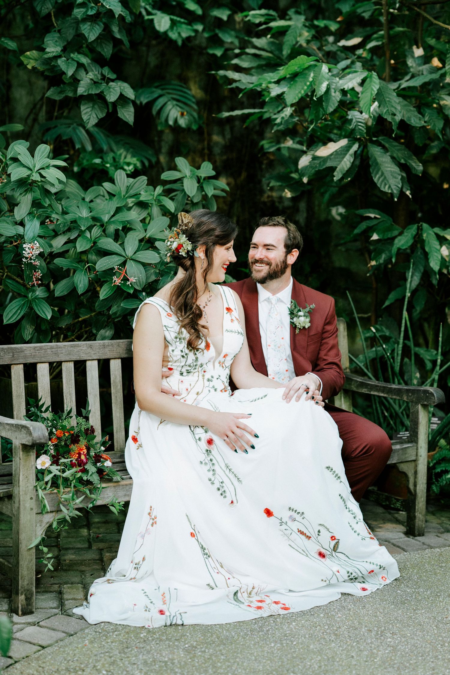 Romantic wedding couple sharing a joyful moment on a rustic garden bench surrounded by vibrant foliage.