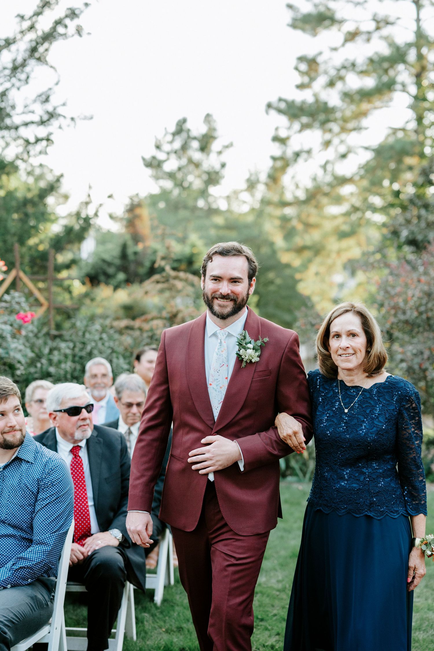 People gather at an outdoor wedding ceremony with pine trees and a gentleman walking down the aisle in a burgundy suit.