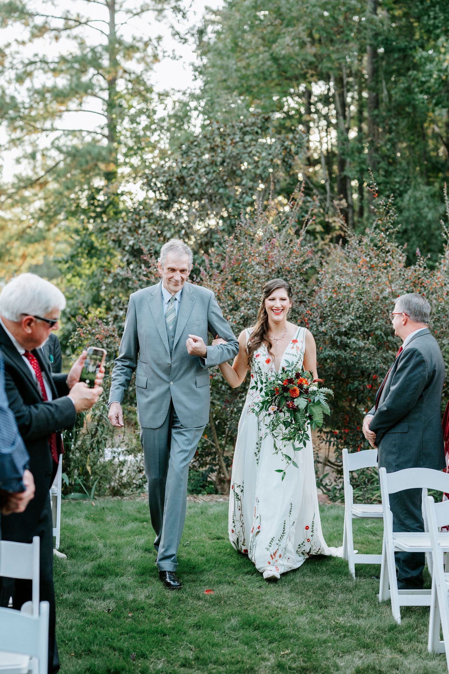 A wedding processional moment on an outdoor ceremony lawn with white chairs and autumn foliage.