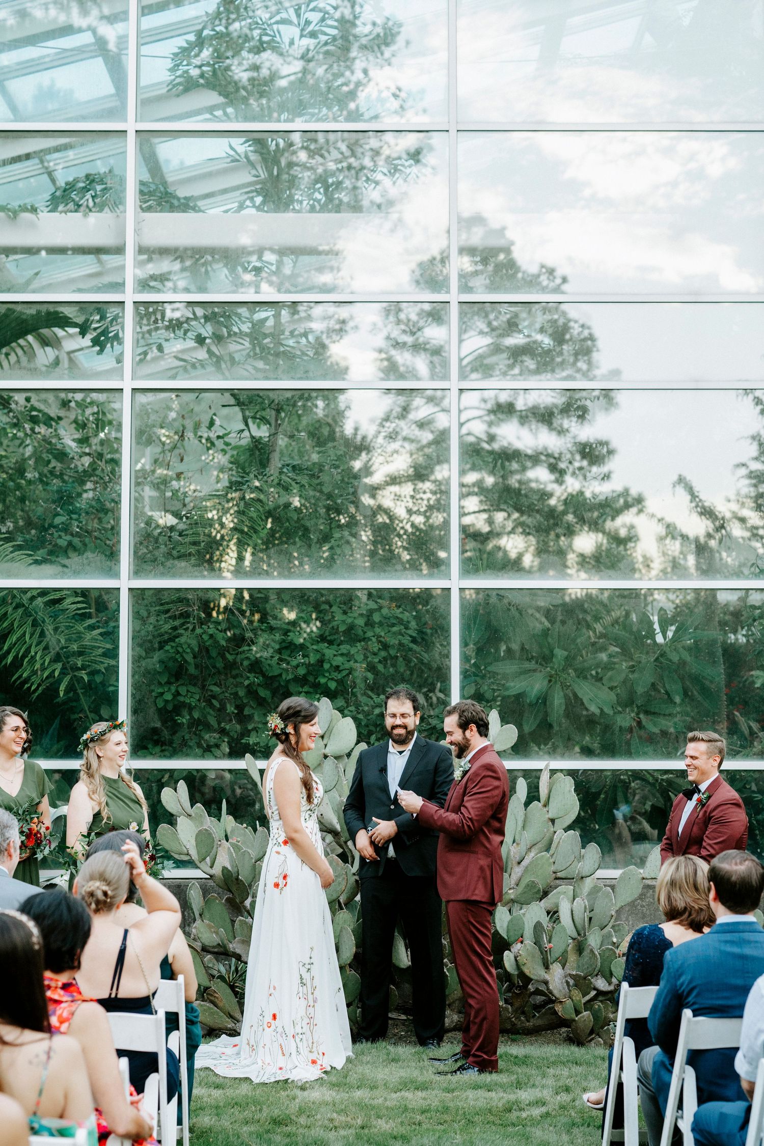 Wedding guests celebrate during an outdoor ceremony at a glass-enclosed greenhouse venue with lush greenery.