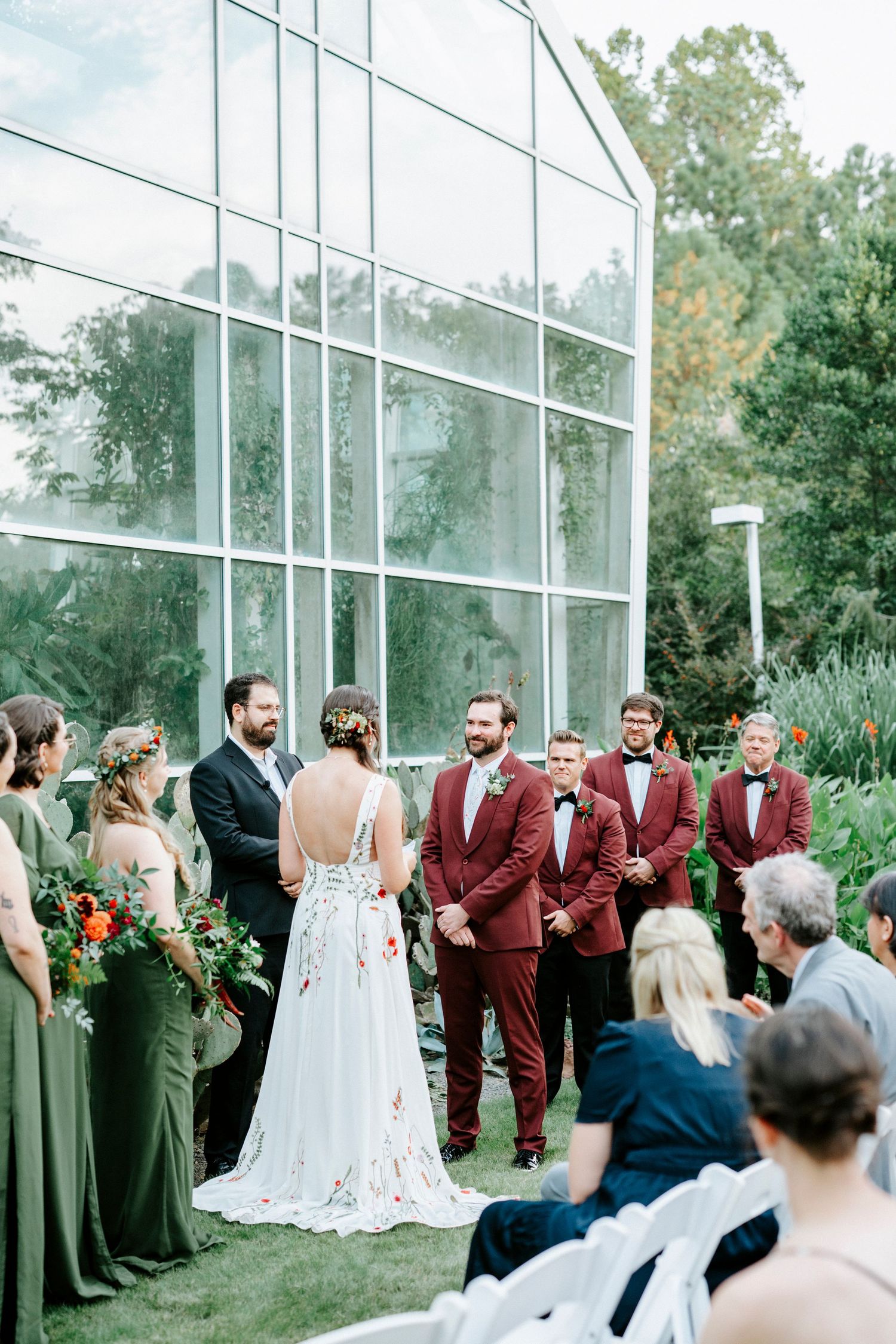 Wedding party in burgundy and sage green attire gathers in front of glass wall for formal portraits.
