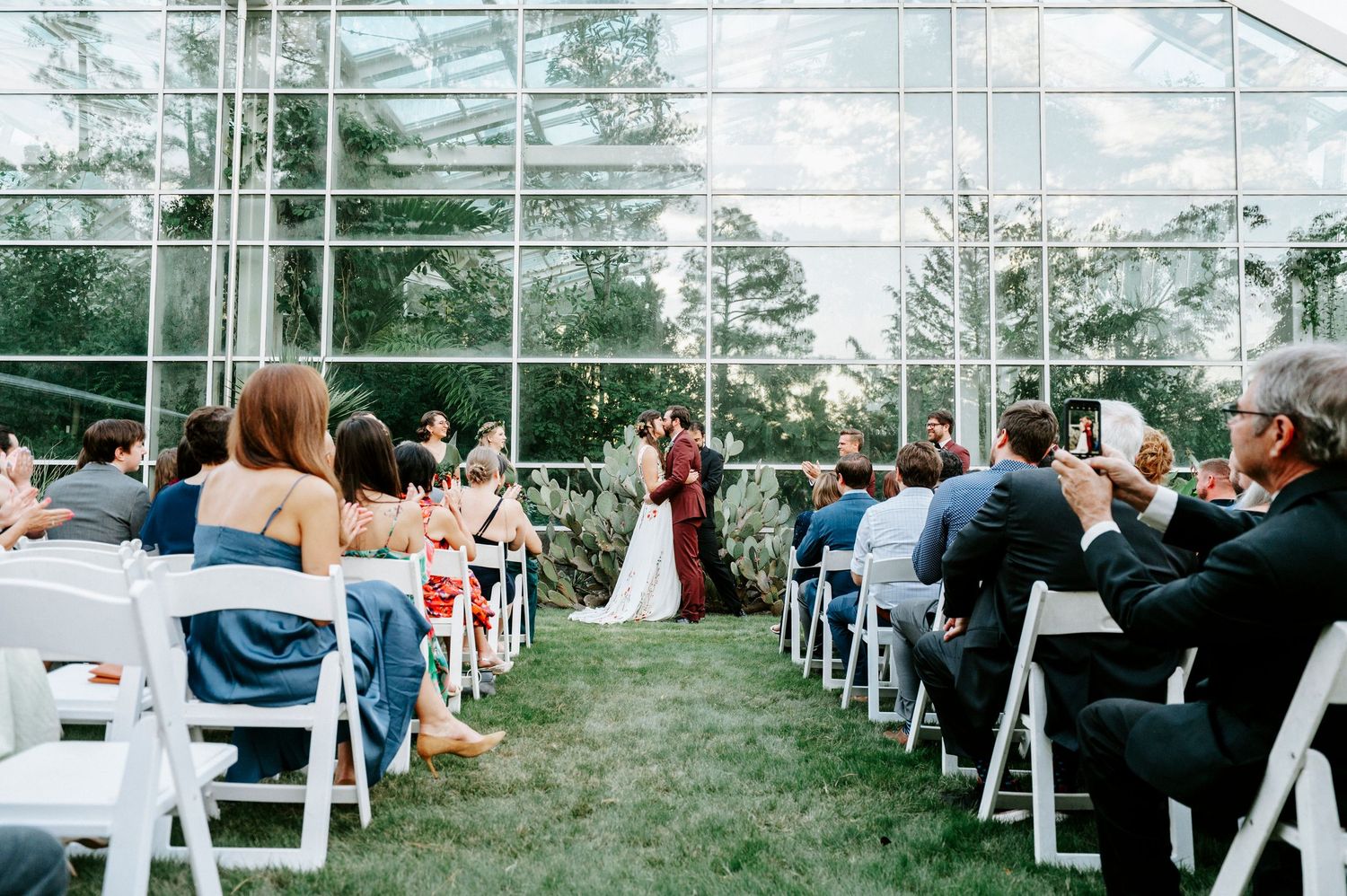 Indoor wedding ceremony in a glass-enclosed venue with white chairs and guests taking photos of the couple.