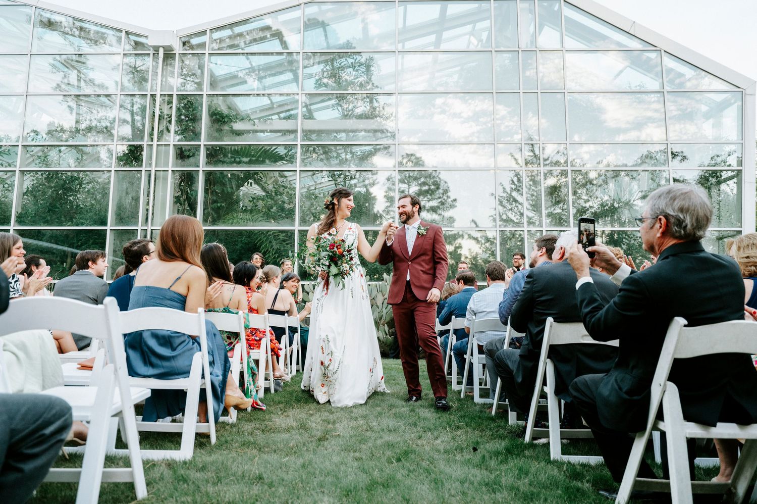 Wedding ceremony setup inside a glass greenhouse with white chairs and natural lighting.