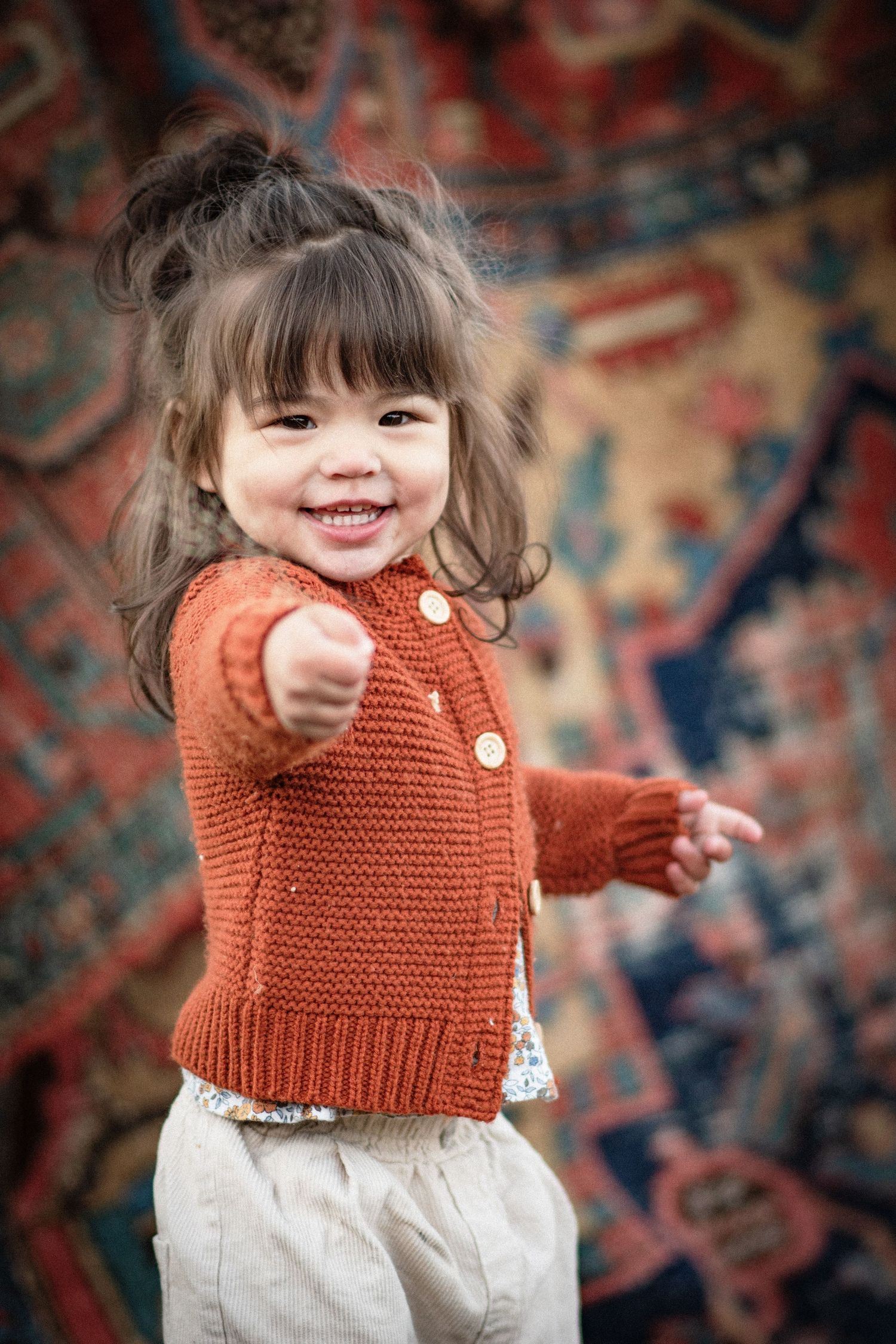 Adorable smiling toddler girl in rust orange knit cardigan reaching out toward camera indoors. Photo by Xenophon Photography in Maine.
