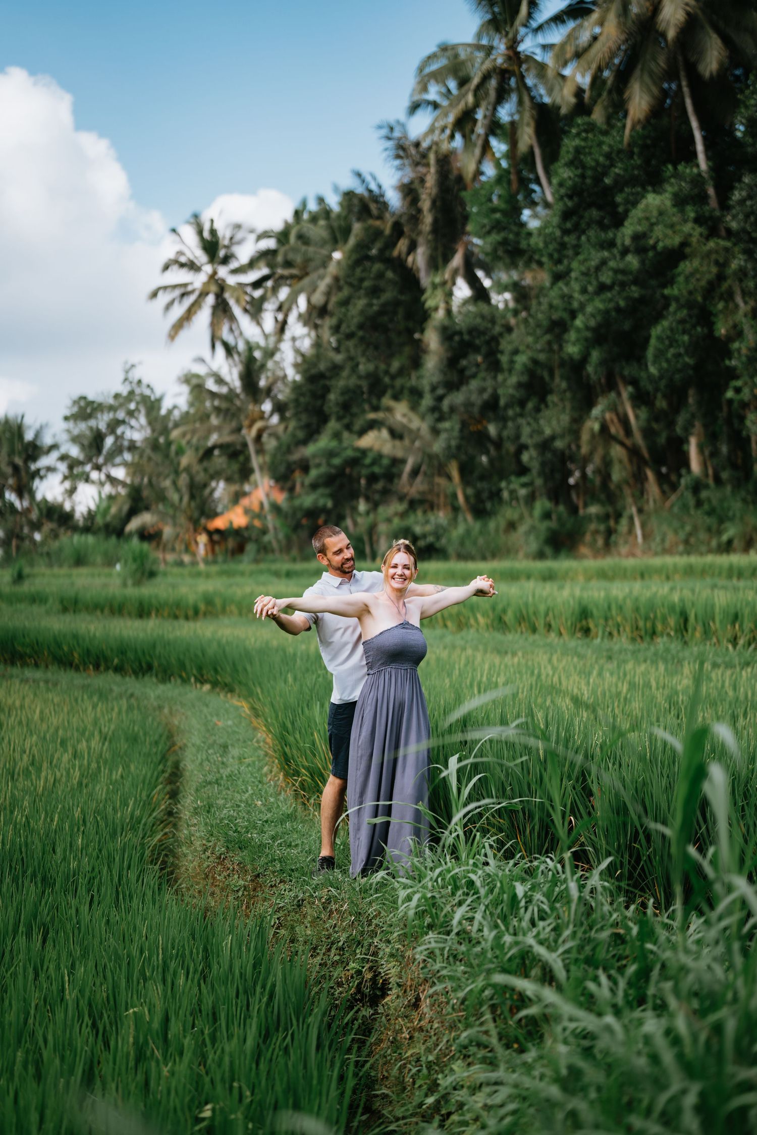 Two people pose playfully in a vibrant green rice field with palm trees in the background.