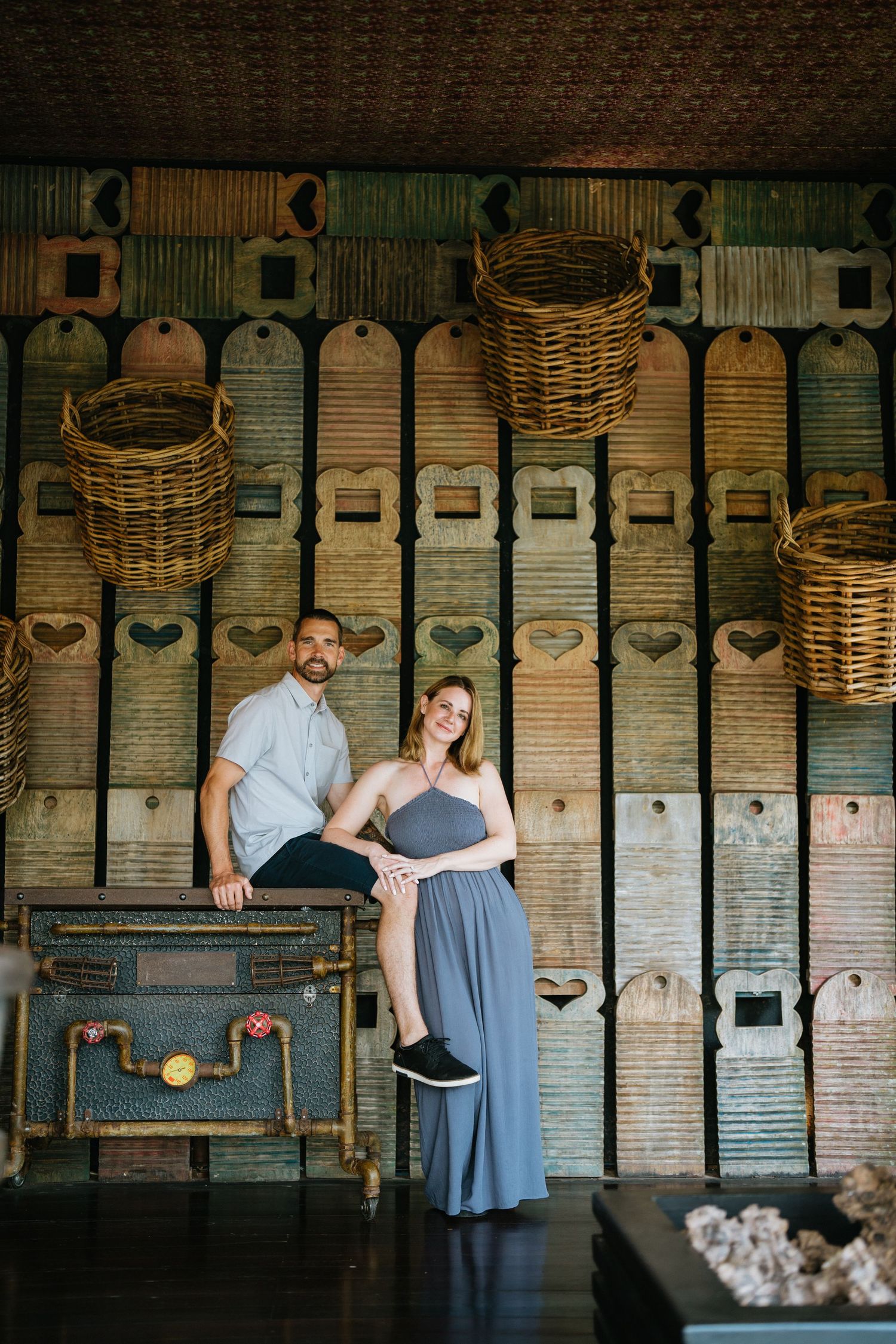 Two people sit together on a rustic bench with woven baskets hanging overhead.
