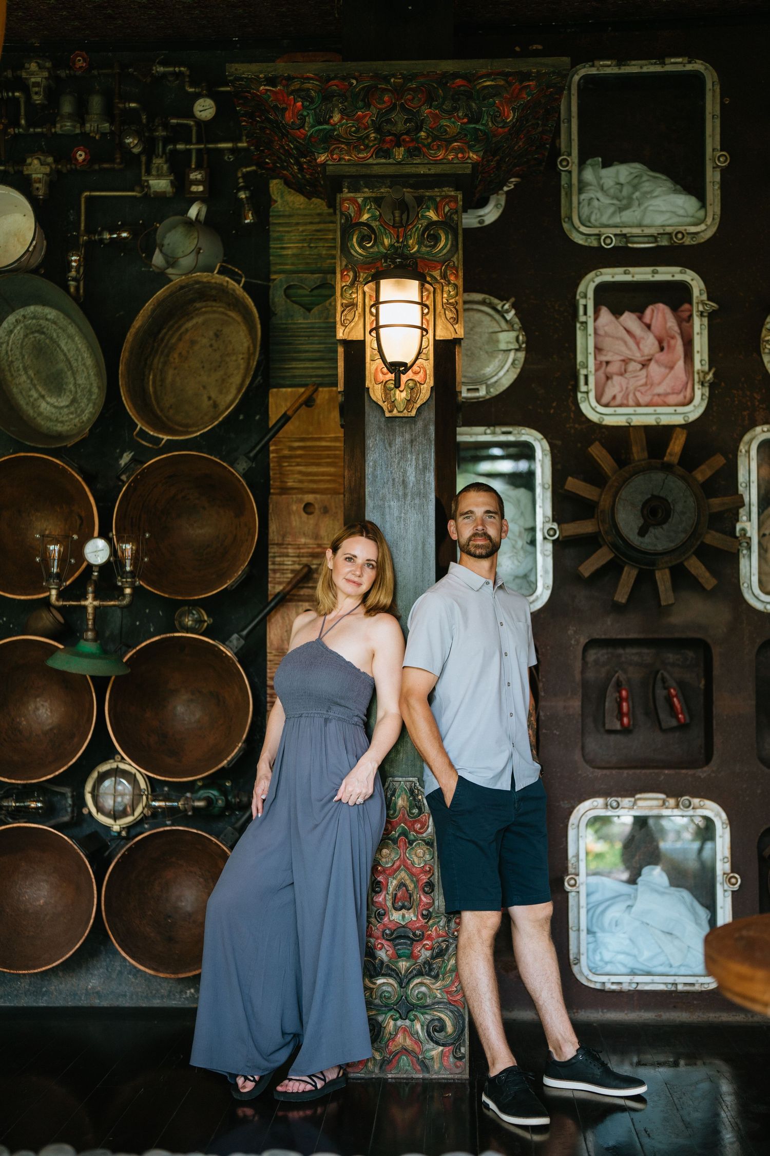 Couple poses against industrial wall decorated with vintage metal gauges and machinery in moody lighting.