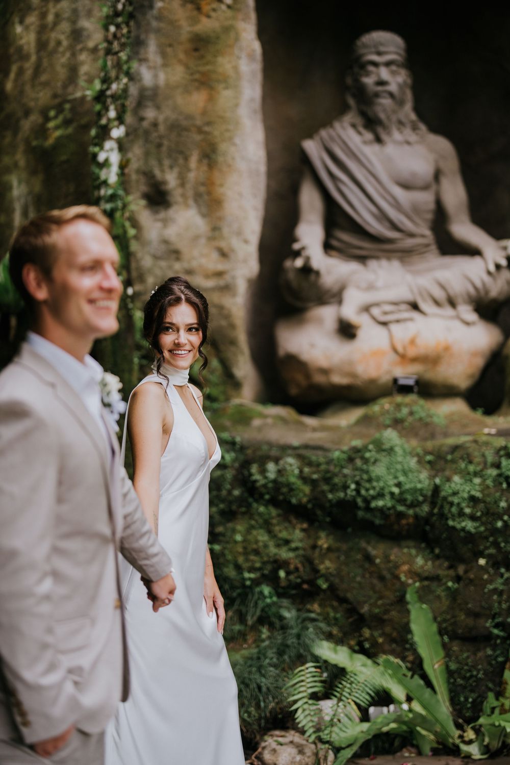 Wedding couple poses in front of stone sculptures in a garden setting.