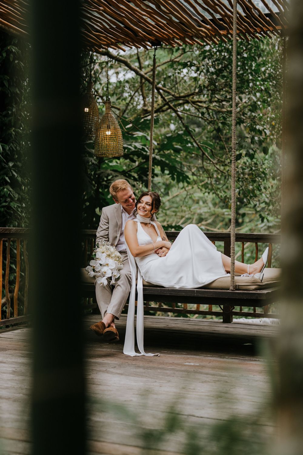 Wedding couple relaxes on wooden deck surrounded by tropical foliage.
