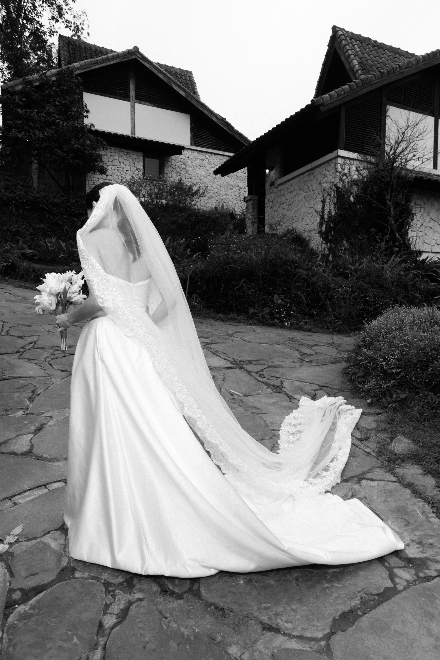 Black and white rear view of bride in elegant gown with long veil on stone path.