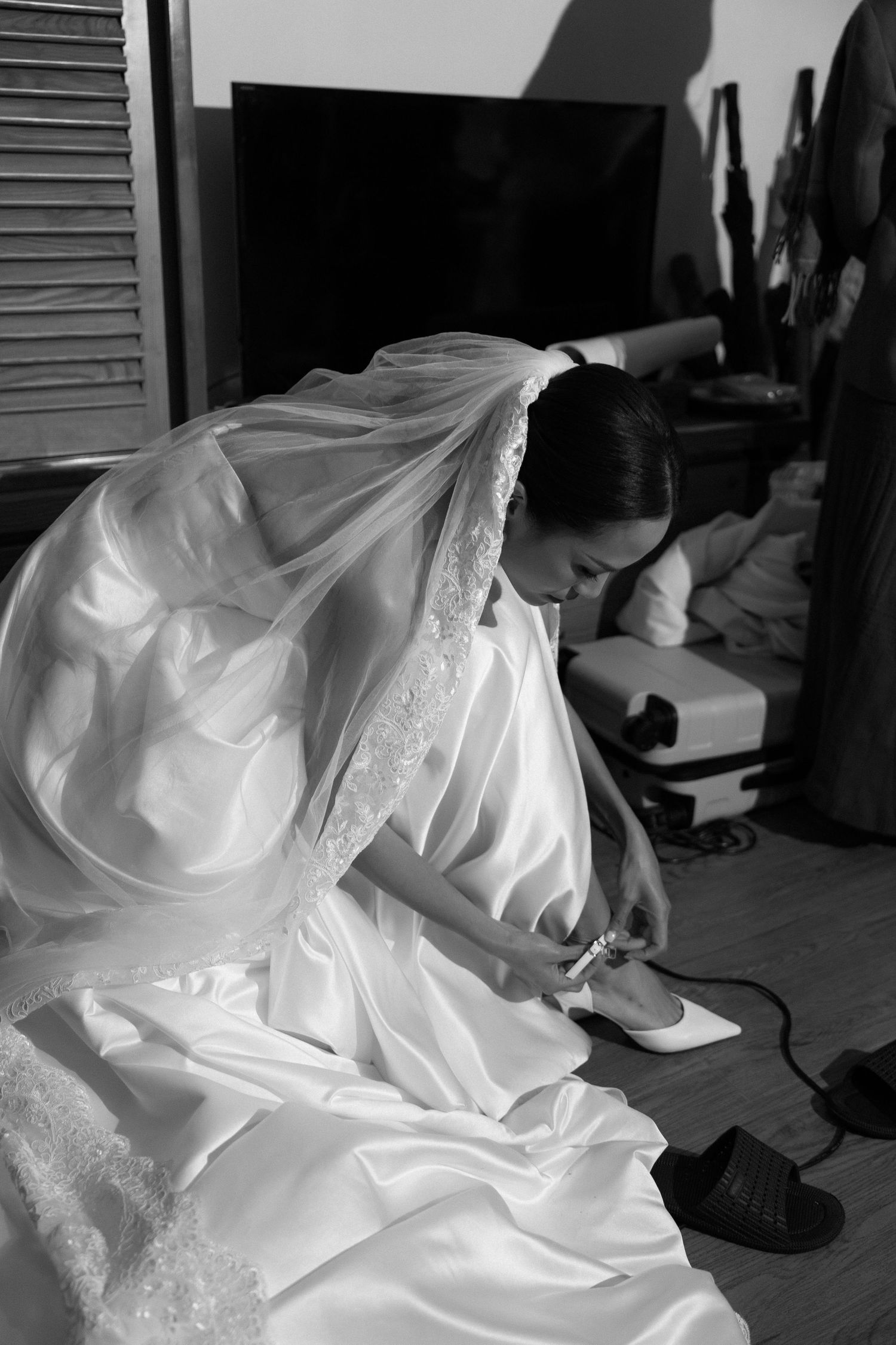 Black and white photo of bride in wedding dress putting on heels indoors.