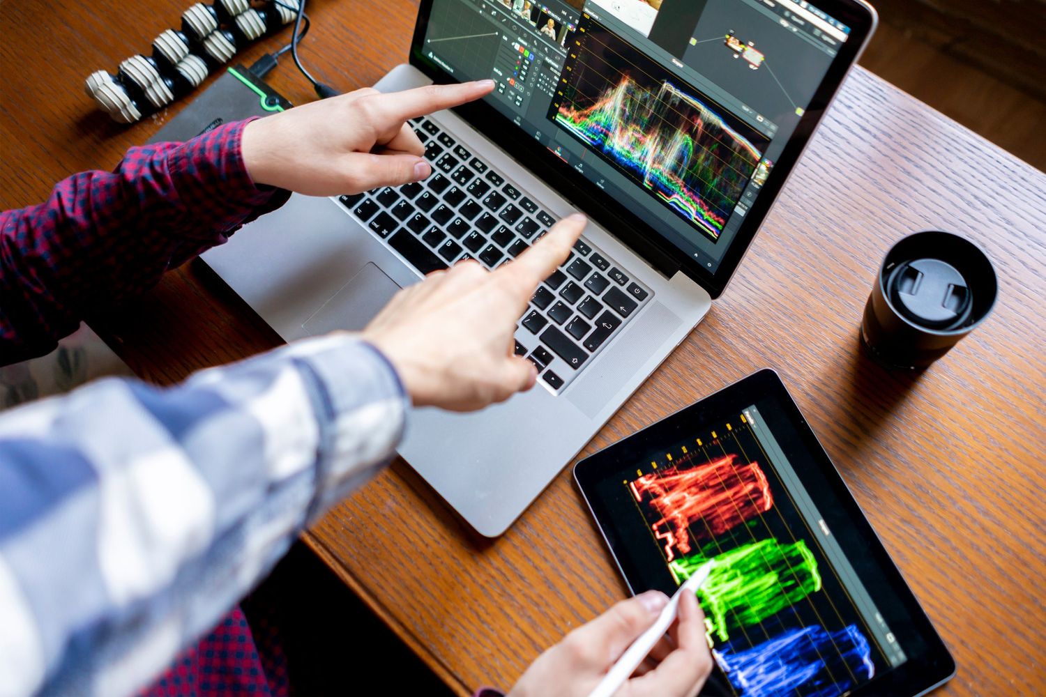 Hands working on laptop and tablet with photo editing software and coffee on wooden desk.