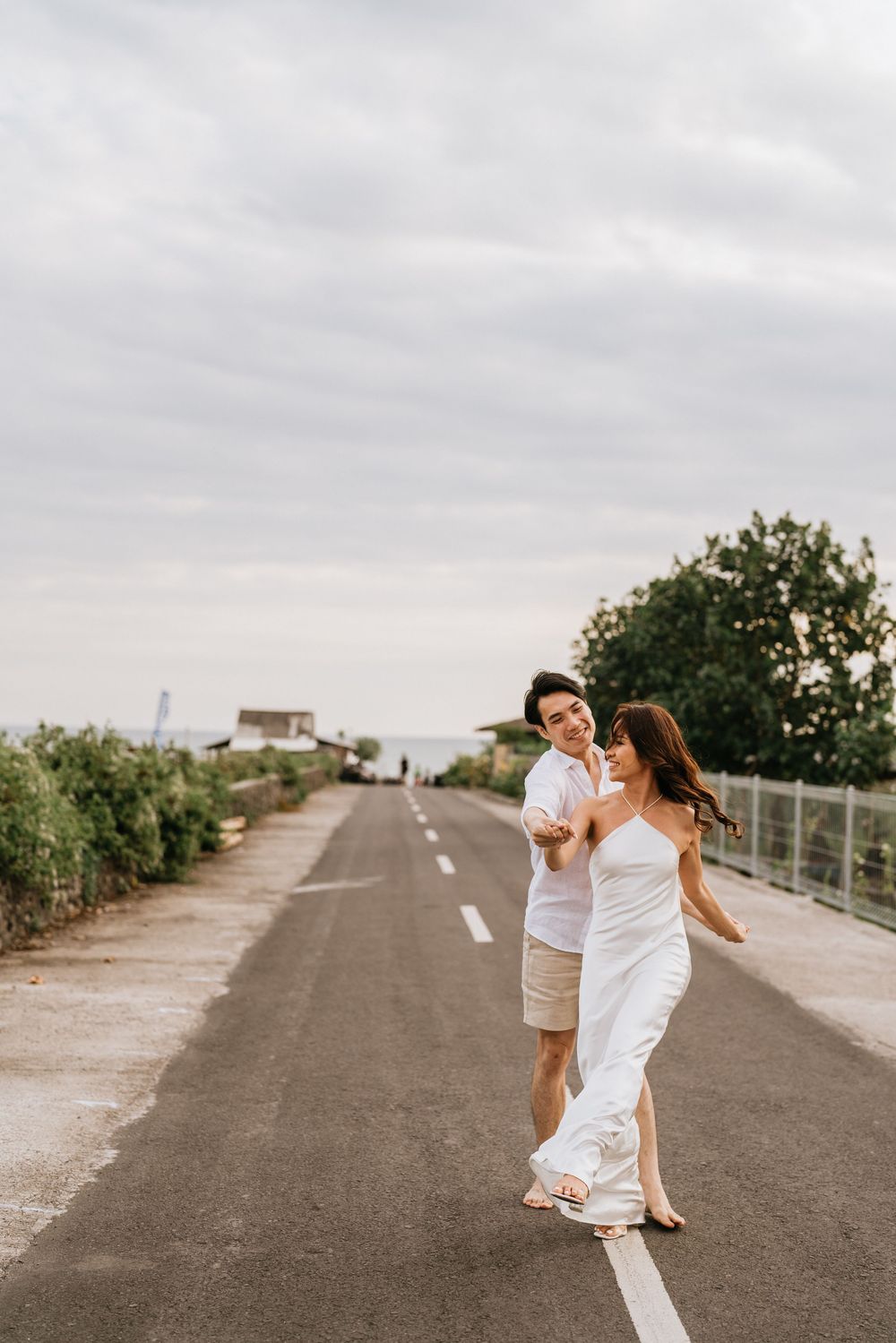 A couple dressed in white runs playfully down a path surrounded by greenery.