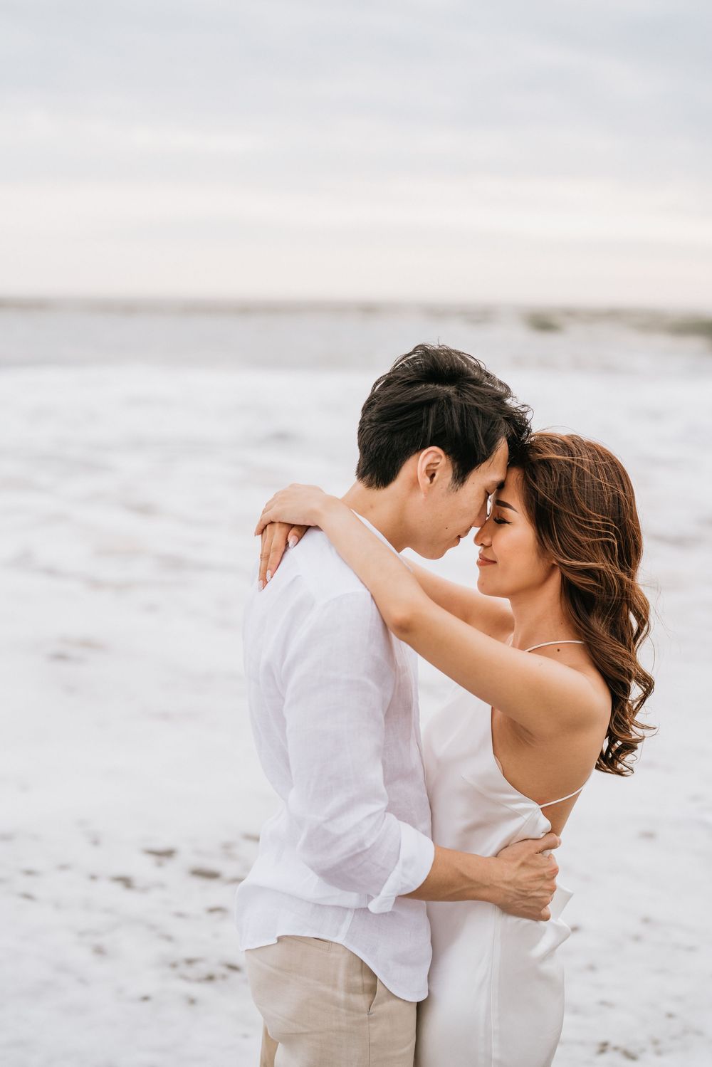 A couple shares an intimate embrace on a sandy beach with waves in the background.