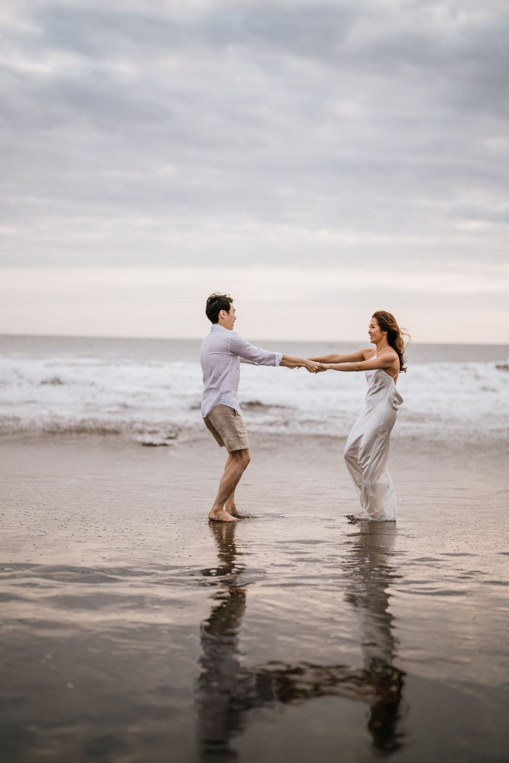 A couple splashes and plays in shallow ocean water during an overcast day.