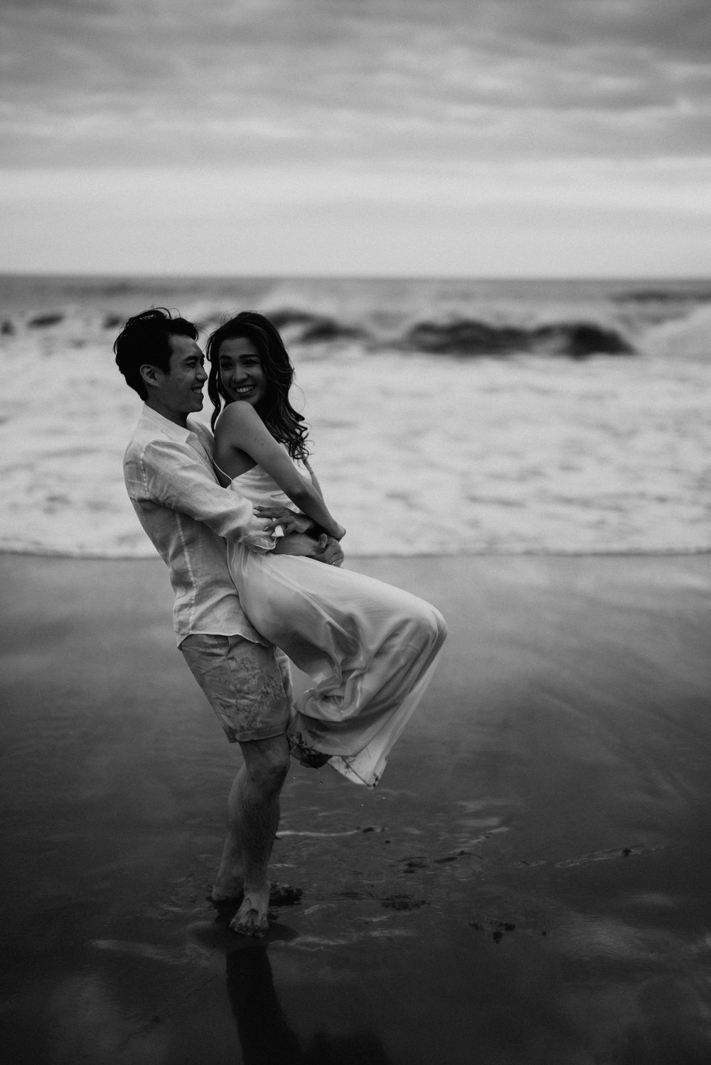 A dramatic black and white photo shows a couple embracing on a moody beach.