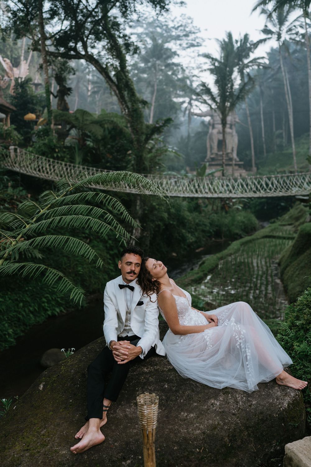 Artistic wedding photo on a rustic bridge amidst lush jungle vegetation.