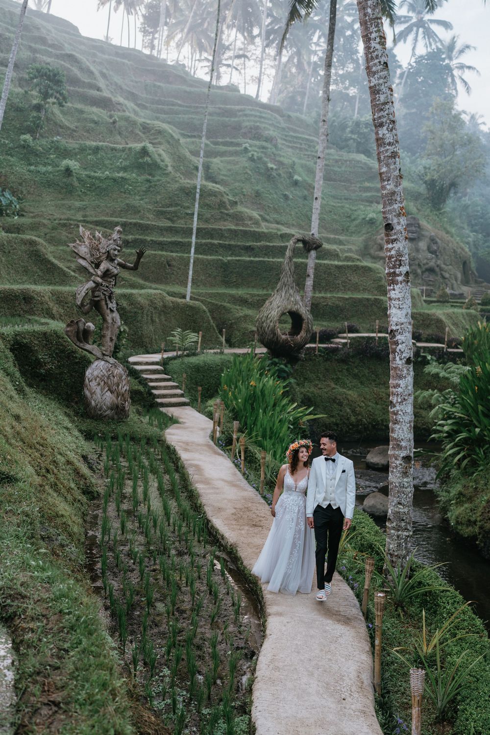 A couple walks along a winding path through lush terraced rice fields and palm trees in Bali.