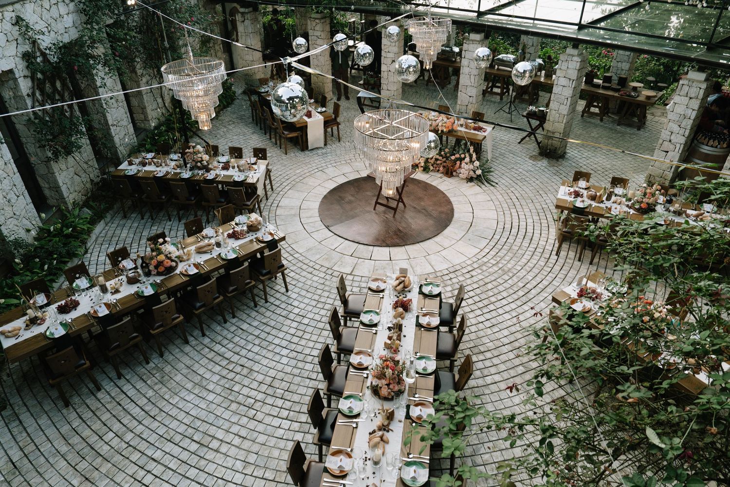 Aerial view of elegant outdoor wedding reception with circular paved courtyard, long decorated tables, and chandelier lighting.