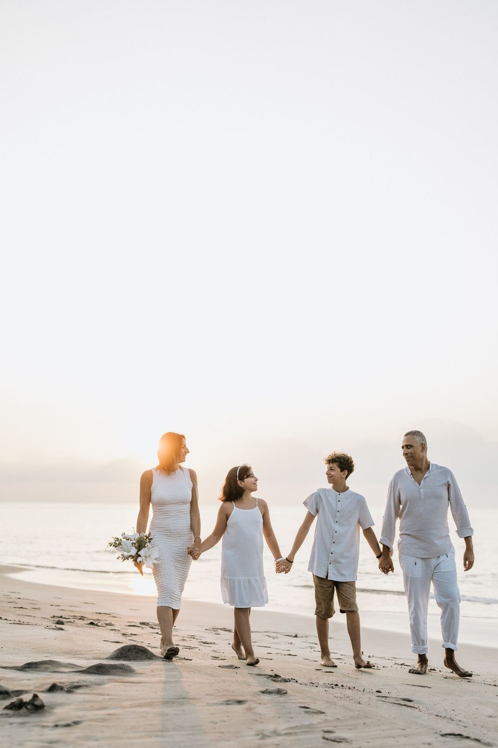 Family walking together on a beach wearing coordinated white outfits at sunset.