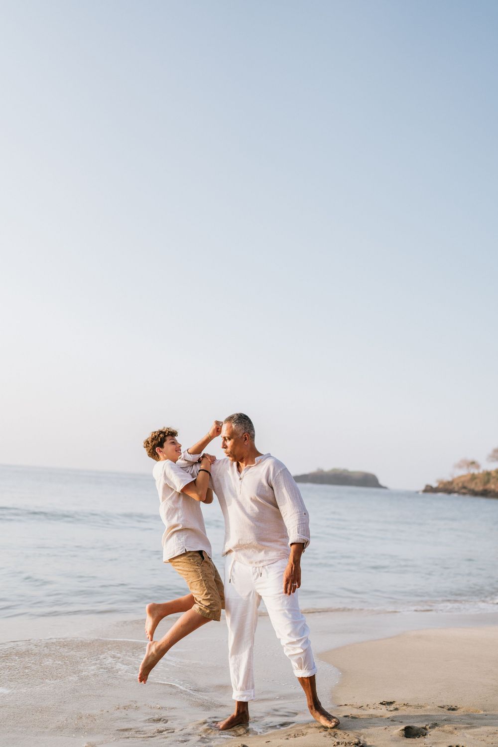 Playful family moment captured on beach with ocean waves and rocky coast in background.