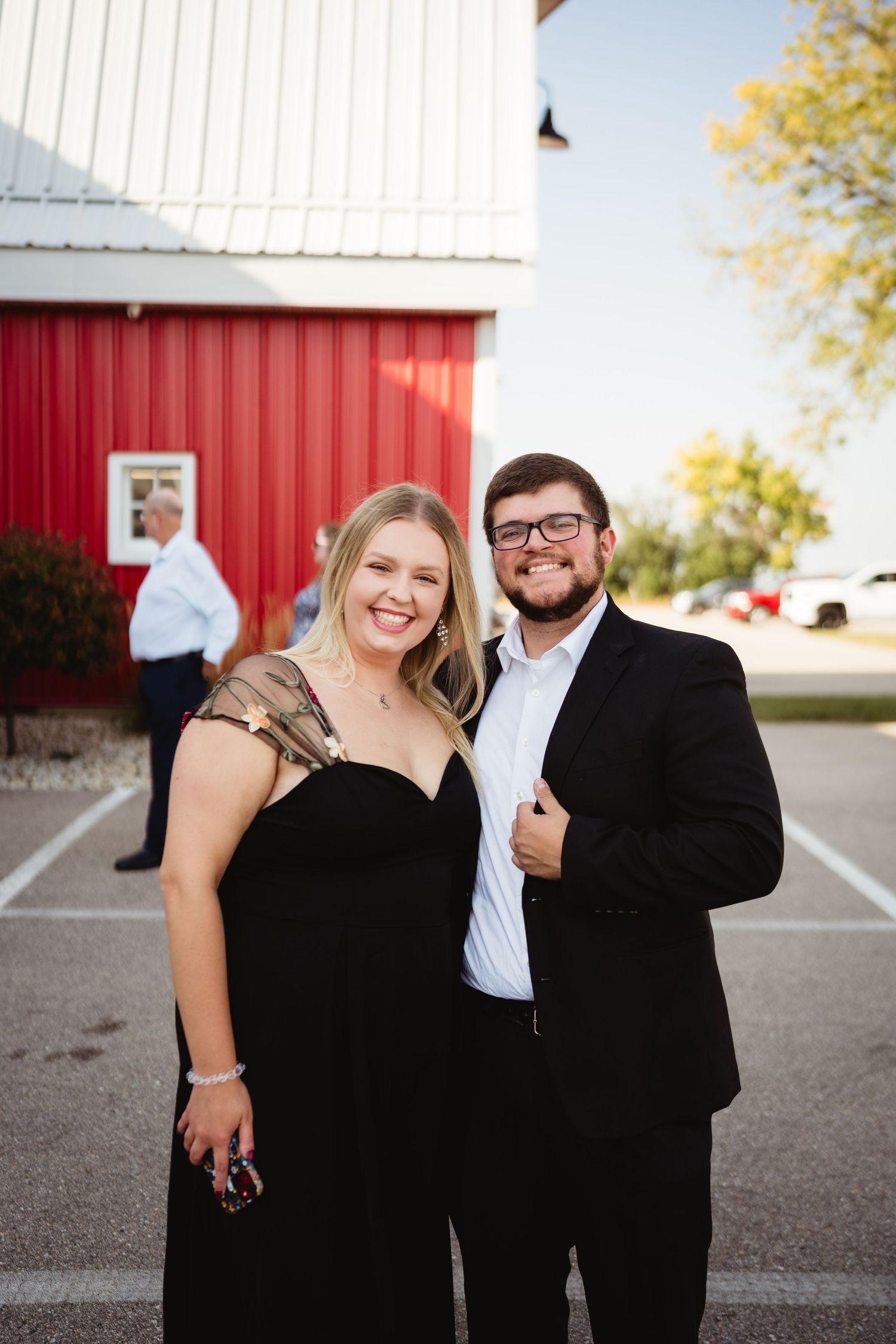 A couple dressed in formal black attire poses outdoors near a red barn wall in a casual photo series.